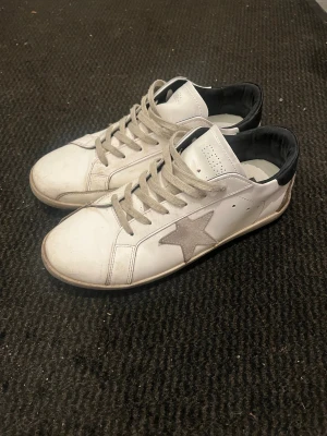 Vita sneakers från Golden Goose - Snygga sneakers från Golden Goose med grå stjärndetalj på sidan, beige skosnören och svart hälparti. Skorna är i storlek 45 och har två slitna hål men är annars i ett tillfredställande skick! 