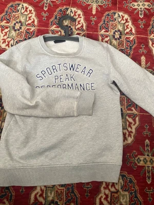 Grå sweatshirt Peak Performance - Snygg grå sweatshirt från Peak Performance med texten 'SPORTSWEAR PEAK PERFORMANCE' broderad framtill. Klassisk rund halsringning och långa ärmar. Perfekt till jeans eller träningsbyxor för en avslappnad look.