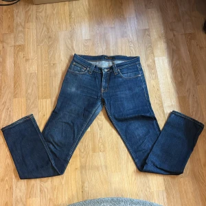 Blå jeans från Nudie Jeans - Säljer ett par klassiska blå jeans från Nudie Jeans med raka ben och snygga kontrastsömmar. Jeansen har fem fickor och coola broderade detaljer på bakfickorna. Materialet är denim i bomull och passformen är avslappnad. Passar slimfit 