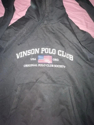 Svart hoodie Vinson Polo Club S - Svart hoodie från Vinson Polo Club med tryck framtill och amerikansk flagga. Klassisk känguruficka och dragsko i huvan. Perfekt för en avslappnad och sportig stil.