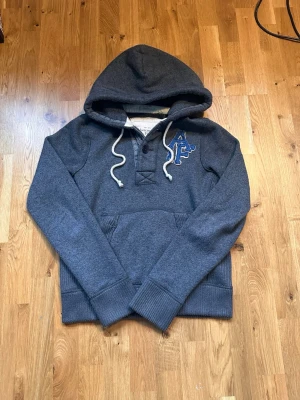 Mörkgrå hoodie från Abercrombie & Fitch - Säljer en mörkgrå hoodie med broderad A&F-logga i blått och vitt på bröstet. Hoodien har huva med vita snören, knappslå framtill och en stor magficka. Materialet är mjukt och bekvämt, perfekt för chill dagar.