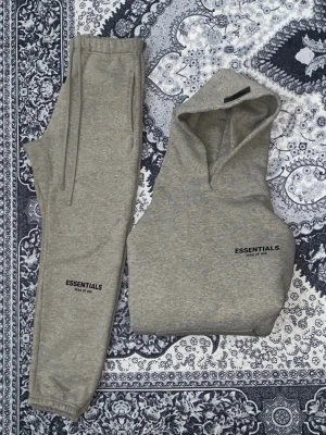 Grå Essentials Tracksuit  - Säljer ett grått loungewear-set från Fear of God Essentials. Setet består av en hoodie med huva och matchande mjukisbyxor, båda med Essentials-logga tryckt i svart. Tillverkad i mjukt bomullsmaterial med ribbade muddar och dragsko i midjan på byxorna. Perfekt för chill dagar hemma.