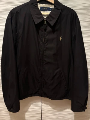 Svart jacka från Polo Ralph Lauren - Stilren svart jacka från Polo Ralph Lauren med klassisk krage och dragkedja framtill. Jackan har två sidfickor och den ikoniska broderade loggan på bröstet. Perfekt för dig som gillar enkel och tidlös design. Storlek LARGE