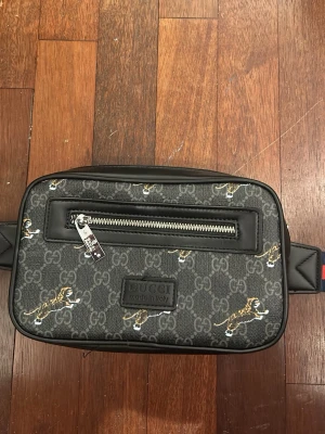 Gucci magväska med tigerprint - Snygg magväska från Gucci i svart med klassiskt GG-mönster och coola tigerdetaljer. Väskan har en silverfärgad dragkedja framtill och justerbart band. Tillverkad i slitstarkt material med läderdetaljer och rymligt innerfack. Perfekt för dig som vill ha både stil och funktion.