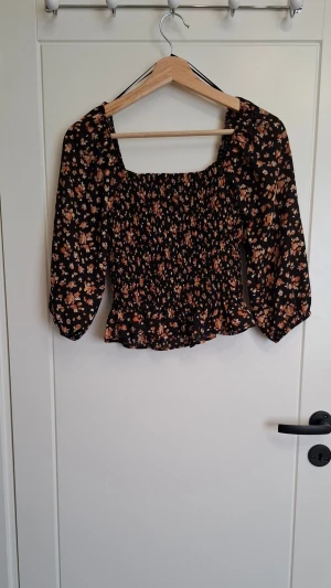 Blommig topp från Paper Crane - Supersöt blommig topp från Paper Crane med smockad kropp och trekvartsärmar. Svart botten med små blommor i orange, beige och vitt. Kvadratisk ringning och volangkant nedtill ger en romantisk vibe. Perfekt att styla med jeans eller kjol!