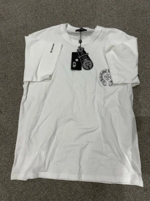 Vit t-shirt från Chrome Hearts - Säljer en clean vit t-shirt från Chrome Hearts med svart logga och emblem tryckt på bröstet. T-shirten har korta ärmar och rund hals. Storleken är M men är lite större i storlek så den psssar L också. Materialet är mjuk bomull och passformen är normal. Perfekt för dig som gillar streetwear och stilrena plagg. Skriv över det minsta lilla eller över den minsta funderingen!🔥🔥