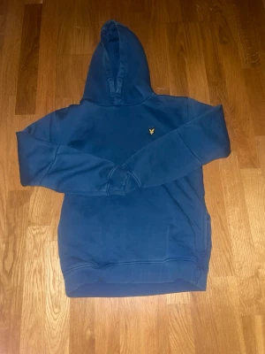 Blå hoodie från Lyle & Scott - Snygg blå hoodie från Lyle & Scott med klassisk huva och det ikoniska gula örnloggan broderad på bröstet. Tillverkad i mjuk bomullsmix som känns skön mot huden. Perfekt för en avslappnad och stilren look.