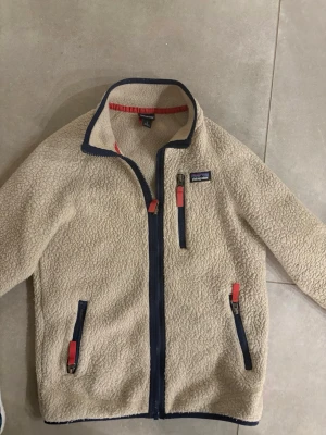 Beige fleecejacka från Patagonia - Säljer en beige fleecejacka från Patagonia med marinblå dragkedjor och röda detaljer. Jackan har hög krage, tre fickor med dragkedja och är gjord i mjukt teddyfleece-material. Perfekt för lager-på-lager och riktigt skön att ha på sig.