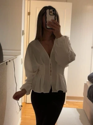 Vit v-ringad blus med knappar - Stilren vit blus med v-ringning och dekorativa knappar framtill. Blusen har lång ärm med lätt puff och markerad midja som ger en feminin siluett. Perfekt att matcha med både jeans och kjol för en trendig look. Storlek 32 men skulle säga mer som 34/36. Endast använd en gång!! SLUTSÅLD PÅ HEMSIDAN