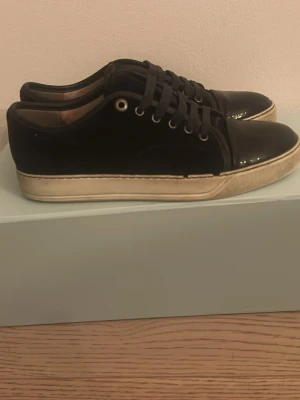 Svarta lackade sneakers från Lanvin - Stilrena svarta sneakers från Lanvin med blank lackad yta och vita sulor. Skorna har svarta skosnören, metallfärgade öljetter och en klassisk låg profil. Perfekta för dig som gillar en clean och modern look.