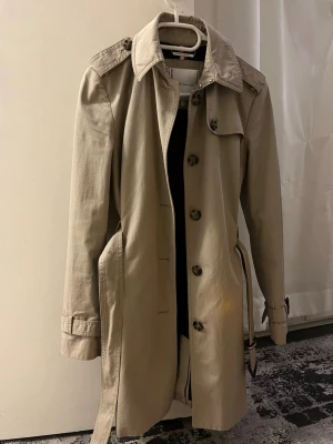Beige trenchcoat från Tommy Hilfiger - Snygg beige trenchcoat från Tommy Hilfiger med klassisk krage, axelklaffar och bälte i midjan. Jackan har stora knappar framtill och på axlarna, samt justerbara remmar vid ärmsluten. Perfekt för dig som vill ha en stilren och tidlös look.