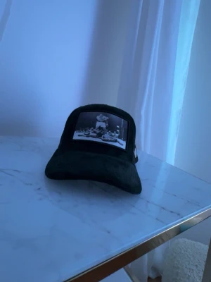 Svart meshkeps med fototryck - Säljer en svart keps med mesh på sidorna och ett stort fototryck framtill som föreställer en klassisk boxningsscen. På sidan sitter ett metallfärgat W-emblem. Kepsen har böjd skärm och justerbar snapback. Perfekt för dig som gillar streetwear och sportiga vibes.