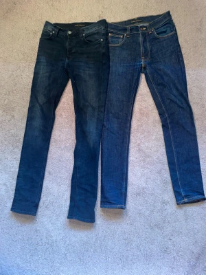 Två par blå jeans från Nudie Jeans - Postar samma dag,Pris kan diskuteras          Säljer två par klassiska blå jeans från Nudie Jeans. Ena paret är mörkblått med slim fit passform och det andra är mellanblått med kontrastsömmar och bakfickor med signaturbrodyr. Båda har fem fickor och knappgylf. Perfekta basjeans för en avslappnad stil. Båda jeansen har en slim fit passform. De mörka paret åt vänster är i ett väldigt bra skick, de åt vänster har ett litet hål så knappast syns, men går att laga gratis hos nudiejeans butikerna. 