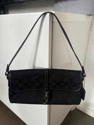 Unik Svart axelväska från Coach. ÄKTA - Snygg svart axelväska från Coach med klassiskt monogrammönster, silverfärgat spänne och detaljer i läder. Väskan har en kort axelrem och en tag med Coach-logga. Perfekt för dig som gillar stilrena accessoarer. Äkta. Använd gärna köp nu🫶