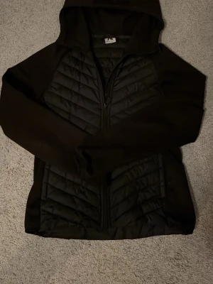Svart pufferjacka från McKinley - Snygg svart pufferjacka från McKinley med huva och dragkedja framtill. Jackan har quiltade partier på bröstet och magen samt släta ärmar. Perfekt för kalla dagar och enkel att matcha med olika outfits.
