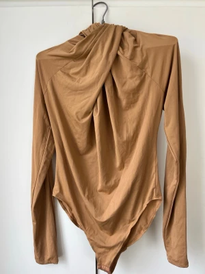 Beige långärmad body från H&M - Säljer en beige långärmad body från H&M med draperad detalj framtill. Toppen har en kroppsnära passform och med knäppning i grenen. Perfekt att styla med jeans eller kjol för en trendig look.