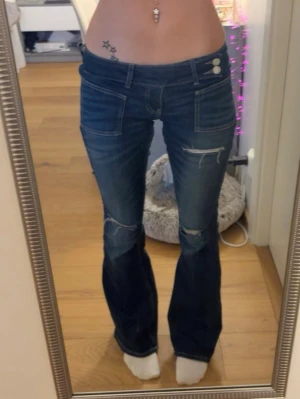 Blå bootcut jeans från Gina Tricot - Säljer ett par blå bootcut jeans från Gina Tricot i storlek XS. Jeansen har slitna detaljer vid knäna och låren, två knappar i midjan och stora fickor fram.