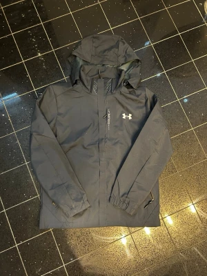 Svart vindjacka från Under Armour - Snygg svart vindjacka från Under Armour med huva och dragkedja framtill. Jackan har ett diskret tryckt logotyp på bröstet och texten 'Explore' vid dragkedjan. Tillverkad i ett lätt och vattenavvisande material, perfekt för blåsiga dagar.