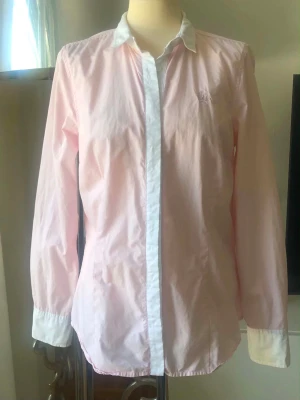Ljusrosa skjorta  - Stilren ljusrosa skjorta med vit krage och knappslå. Skjortan har långa ärmar och diskret broderad logga på bröstet. Perfekt för en clean och klassisk look. Armhåla till armhåla 50 cm. Storlek S/M