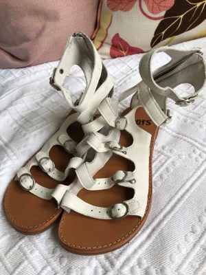 Vita läder gladiatorsandaler från Roots - Säljer ett par vita gladiatorsandaler från Roots med flera remmar och spännen. Sandalerna har dragkedja bak för enkel påtagning och en brun sula i läderlook. Perfekta för dig som gillar en edgy och trendig stil.