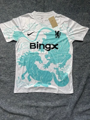 Nike Chelsea tröja  - Snygg Chelsea matchtröja från Nike i vitt med turkost lejon- och hästmotiv över hela framsidan. Tröjan har svart Bingx-logga, klubbmärke och Nike Swoosh på bröstet. Tillverkad i lätt och ventilerande material, perfekt för träning eller match. Finns i S,M och L