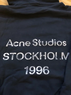 Mörkblå hoodie Acne Studios - Snygg mörkblå hoodie från Acne Studios med stor vit text på ryggen: 'Acne Studios STOCKHOLM 1996'. Klassisk huva och långa ärmar, ribbade muddar och avslappnad passform. Perfekt för dig som gillar streetwear och vill ha något från ett ikoniskt svenskt märke.