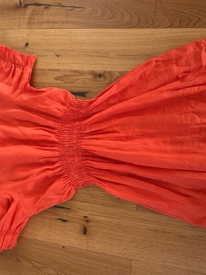 Orange kortklänning från My Aurora - Snygg orange kortklänning från My Aurora i luftig linneblandning. Klänningen har korta ärmar, smock i midjan och en avslappnad passform som ger en cool och bekväm look. Perfekt för varma dagar och sommarens alla äventyr.