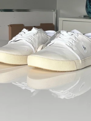 Vita Polo Ralph Lauren sneakers - Snygga vita sneakers från Polo Ralph Lauren med klassisk låg profil och vit logga på sidan. Skorna har vita snören, diskret beige sula och är tillverkade i canvasmaterial. Extra skosnören i off-white följer med. 