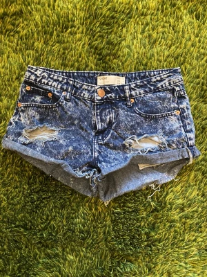 Cotton On Ripped Denim Booty Shorts - Säljer ett par blå jeansshorts från Cotton On med slitna detaljer och upprullade benslyt. Shortsen har klassisk femficksdesign, knappgylf och en cool tvätt. De är för girlypops som vill ha korta shorts och serve cunt.