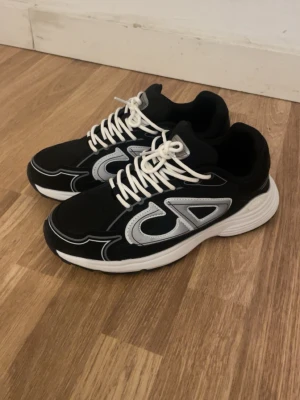Svarta sneakers med vit sula - Snygga svarta sneakers från DAP med vit tjock sula och vita detaljer. Skorna har vita snören och en stor silverfärgad logga på sidan. Ovandelen är i textil och syntet, vilket ger en sportig och modern look.