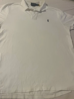 Vit piké t-shirt från Polo Ralph Lauren - Klassisk vit piké t-shirt från Polo Ralph Lauren med korta ärmar och broderad marinblå logga på bröstet. Tröjan har krage och knappar vid halsen. Tillverkad i mjuk bomull som känns skön mot huden.