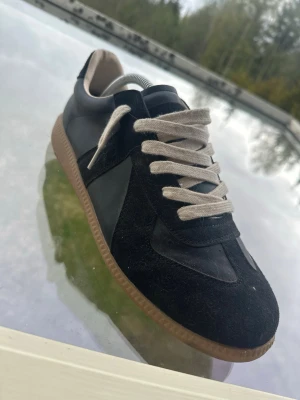 Svarta sneakers med mockadetaljer - Snygga svarta sneakers med beige snörning och bruna gummisulor. Skorna har en mix av mocka och slätt läder på ovandelen, vilket ger en stilren och klassisk look. Perfekta för dig som gillar minimalistisk design och neutrala färger.