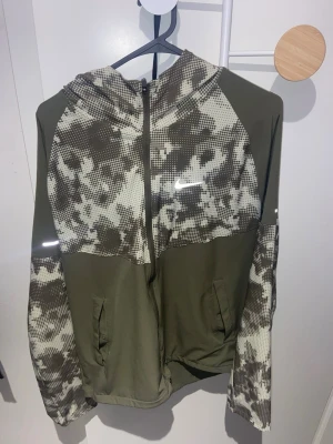 Nike miler Grön Set - Grön tracksuit från Nike med hel dragkedja och camomönstrade partier i vitt och svart på huva, axlar och ärmar. Har två fickor framtill och en liten vit Nike-logga på bröstet. Tillverkad i ett mjukt och stretchigt material, perfekt för en sportig look. Kan tänka mig gå ner i pris vid snabbaffär.