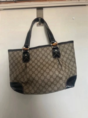 Gucci beige monogram handväska - Snygg beige handväska från Gucci med klassiskt GG-monogram över hela väskan. Väskan har svarta skinndetaljer på handtag och hörn samt guldfärgade metalldetaljer. Perfekt storlek för att få plats med allt du behöver och ger en lyxig touch till din outfit.
