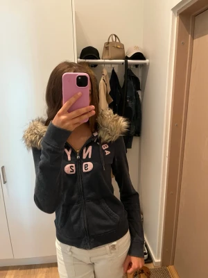 Mörkblå Abercrombie hoodie med fuskpäls - Mysig mörkblå hoodie från Abercrombie med dragkedja, rosa tryck på bröstet och stor huva kantad med fluffig fuskpäls. Två fickor framtill och långa ärmar. Perfekt för dig som gillar en chill och snygg streetstil. Köpt på vinted💗