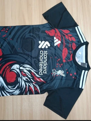 Liverpool FC Adidas svart matchtröja - Snygg svart Liverpool FC matchtröja från Adidas med grafiskt mönster i rött och vitt, samt klubbmärke och sponsortryck på bröstet. Tröjan har korta ärmar och rund hals. Perfekt för dig som älskar fotboll och vill sticka ut på planen.