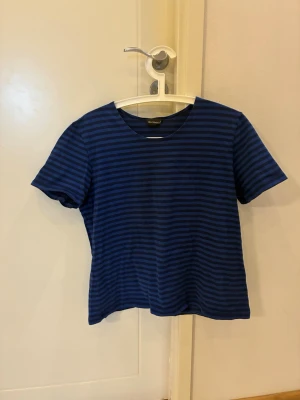 Blå randig t-shirt från Marimekko - Snygg blå t-shirt från Marimekko med tunna mörkblå ränder. Klassisk rund halsringning och korta ärmar. Perfekt för dig som gillar stilrena och tidlösa plagg med en twist. Tillverkad i mjuk bomull som känns skön mot huden.
