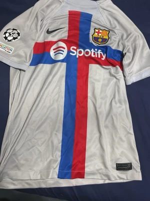 Barcelona bortatröja Nike grå - Snygg grå bortatröja från FC Barcelona med röd och blå korsdesign på framsidan. Tröjan har klubbmärke, Nike-logga och Spotify som sponsortryck. Tillverkad i lätt och ventilerande Dri-FIT-material, perfekt för fotboll eller träning.