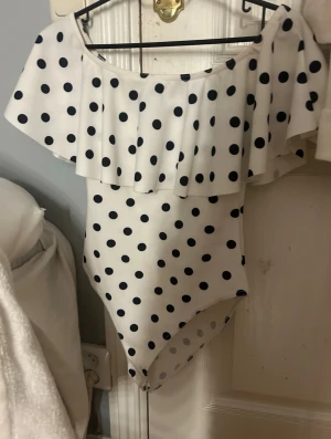 Vit prickig offshoulder body   - Vit offshoulder body med svarta prickar och bred volang över axlarna. Toppen är ärmlös och har en figurnära passform. Tillverkad i stretchigt syntetmaterial som ger en bekväm känsla och trendig look.