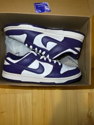 Nike Dunk Low vit/lila sneakers - Nike Dunk Low sneakers i vitt och lila läder med klassisk Swoosh på sidan. Lågt skaft, vita skosnören och perforerad tåbox för extra ventilation. Snygg retrostil som passar perfekt till streetwear och casual outfits. Kontakta mig för fler bilder.