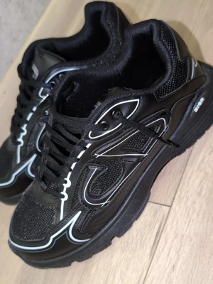 Svarta sneakers med vita detaljer - Stilrena svarta sneakers från Skechers med vita och grå detaljer. Ovandelen är i mesh och syntetmaterial för bra andningsförmåga och komfort. Tjock sula för extra stöd och skön känsla. Klassisk snörning och sportig look som passar till många outfits. Använda max 3 ggr 