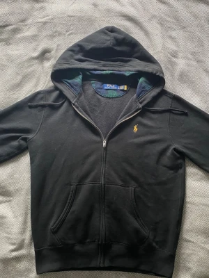 Svart hoodie från Polo Ralph Lauren - Snygg svart hoodie från Polo Ralph Lauren med dragkedja och klassisk gul logga på bröstet. Hoodien har kängurufickor och en fodrad huva med rutigt mönster på insidan. Perfekt för en chill och stilren look.