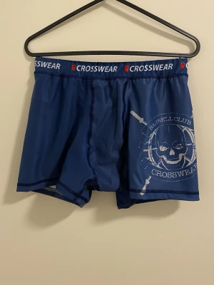 Blå kalsonger från Crosswear - Snygga blå kalsonger från Crosswear med elastisk midjeresår där märket står tryckt. På ena benet finns ett coolt tryck med dödskalle och texten 'Barbell Club Crosswear'. Materialet ser ut att vara mjukt och stretchigt, perfekt för en aktiv livsstil.
