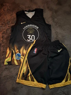Warriors basketset Curry 30 svart/gul - Snyggt basketset från Nike med Golden State Warriors-tema och Curry #30. Setet består av ett svart linne och shorts med gula flames-detaljer, tryckta loggor och nummer. Materialet är lätt och andas, perfekt för basket eller träning.