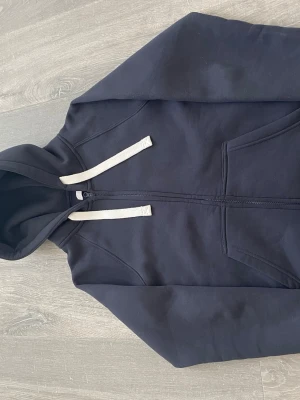 Mörkblå hoodie med dragkedja - Snygg mörkblå hoodie från Russemerch med dragkedja framtill och vita snören i huvan. Hoodien har två fickor på magen och ribbade muddar vid ärmslut och nederkant. Perfekt för en avslappnad och stilren look.
