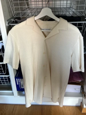 Beige kortärmad skjorta med knappar - Stilren beige skjorta med korta ärmar och klassisk krage. Skjortan har en ribbad struktur och knäpps framtill med diskreta knappar. Perfekt för en avslappnad men snygg look. Endast använd en gång, inga defekter eller fläckar.