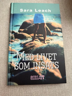 Med livet som insats av Sara Leach - En ungdomsroman med hård pärm, skriven av Sara Leach. Omslaget visar en person vid vatten och klippor, och boken handlar om oväntade äventyr och utmaningar i bergen. Perfekt för dig som gillar spännande berättelser om vänskap och mod.