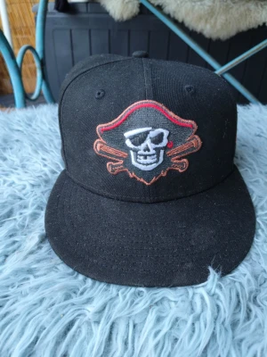 Svart keps med piratmotiv - Säljer en svart keps med platt skärm och broderad dödskalle med pirathatt framtill. På sidan finns ett färgglatt broderi med texten 'Florida State League All-Star Game'. Kepsen har cool streetwear-vibe och passar perfekt för dig som gillar sportiga accessoarer.