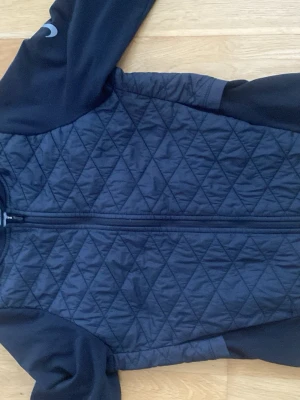 Svart löparjacka från Nike - Snygg svart löparjacka från Nike med quiltat mönster på framsidan och släta ärmar. Jackan har hel dragkedja, hög krage och är tillverkad i funktionsmaterial. Perfekt för löpning eller träning utomhus.