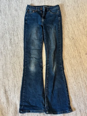 Mörkblå bootcut jeans - Säljer ett par mörkblå bootcut jeans med lätt slitna detaljer framtill och baktill. Jeansen har normal midja och utsvängda ben. Perfekta för dig som gillar retrovibbar och en avslappnad look.